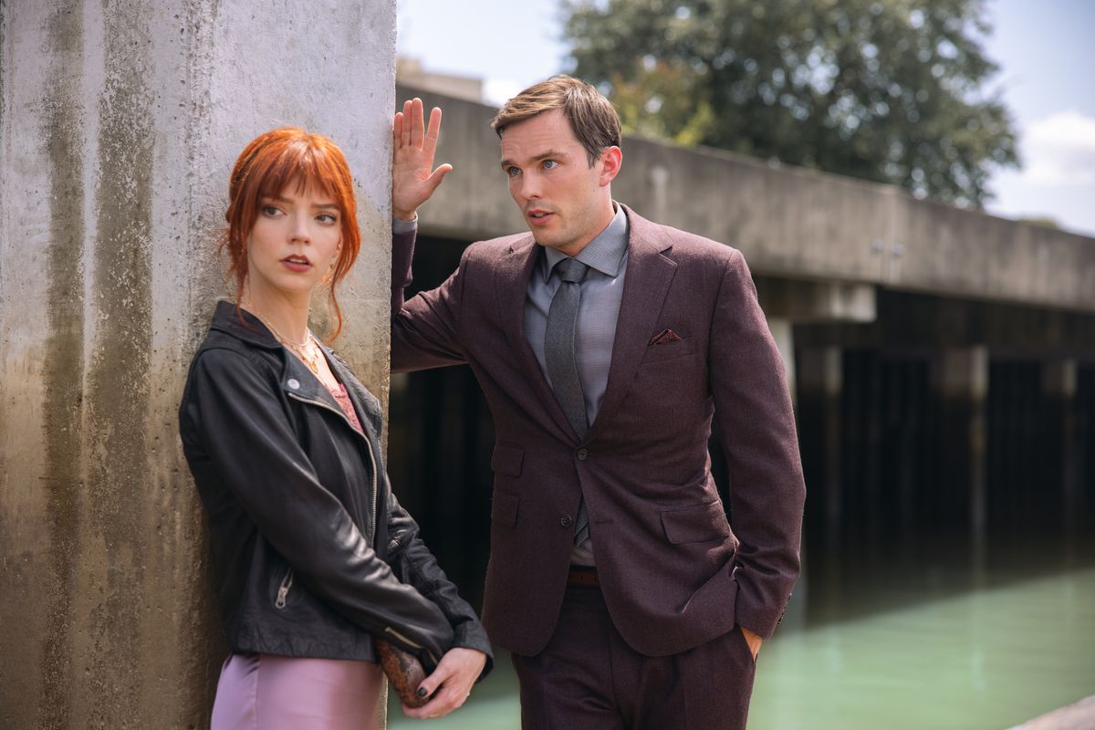 Nicholas Hoult’s Tyler in a suit and tie raises his arm as he speaks to his date Margot (Anya Taylor-Joy) with bright red hair a leather jacket and a purple dress as they stand by a concrete pillar in The Menu