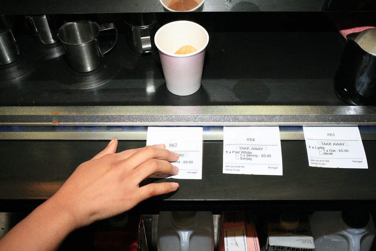 Three restaurant tickets on a metal rail, with a takeaway cup in the foreground.