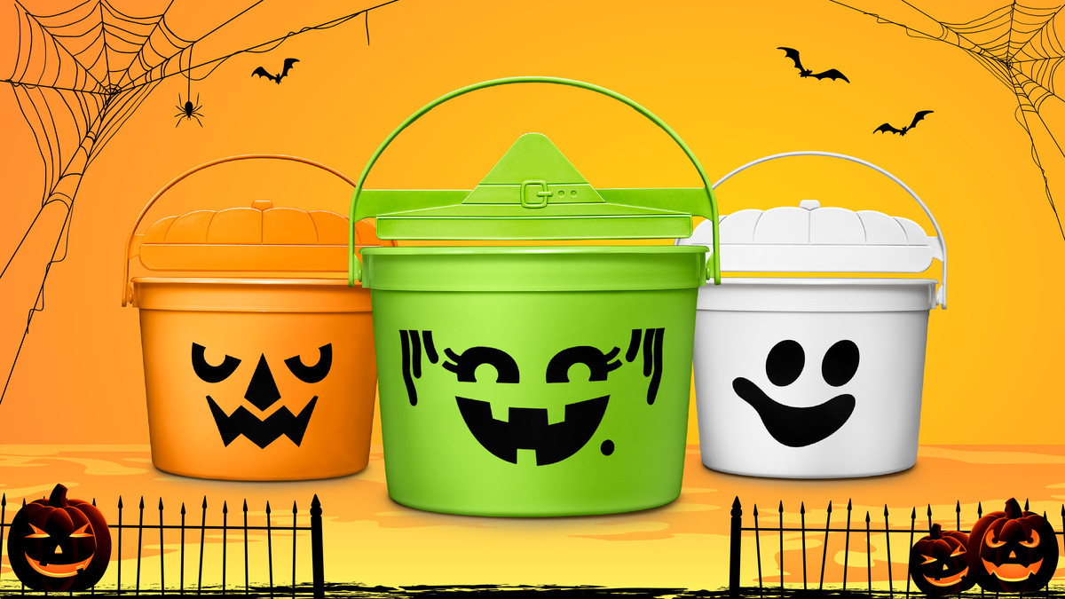 Three plastic buckets, one orange, one green, one white, and all emblazoned with a jack-o-lantern style face. The background is orange with spider webs and bats at the corner