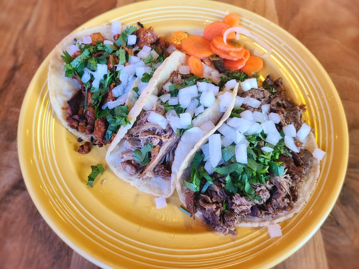 Three tacos are shown on a yellow plate sitting on a wooden table.