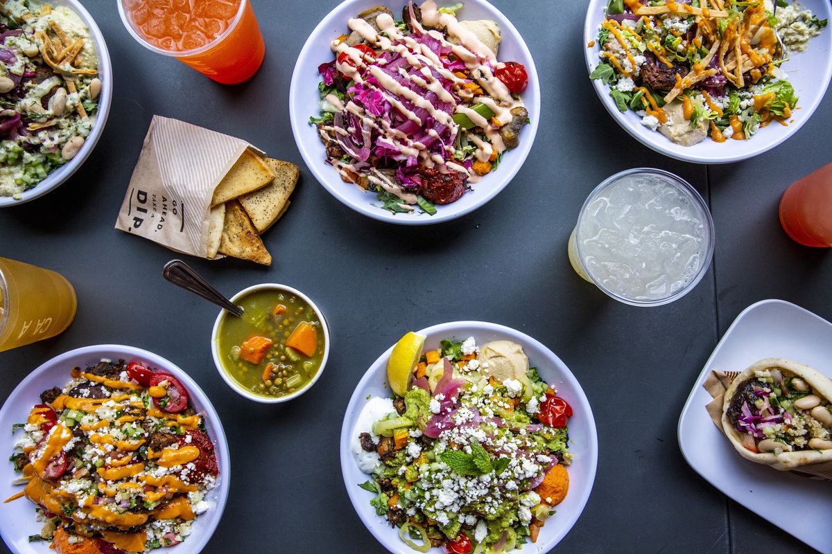 A spread of salads and grain bowls from Cava, arranged on a dark grey background.