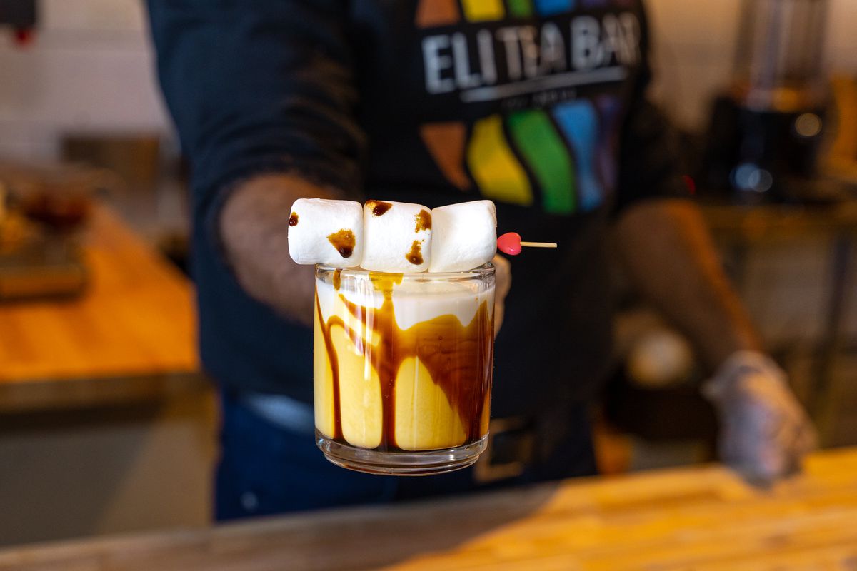 A server holds out a drink in a glass mug with yellow liquid streaked with brown and topped with  three marshmallows on a skewer