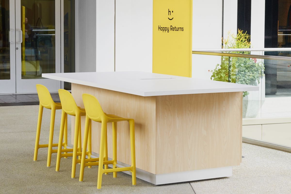 A mock-up of a Happy Returns return desk at a mall has a bare table and three yellow chairs.