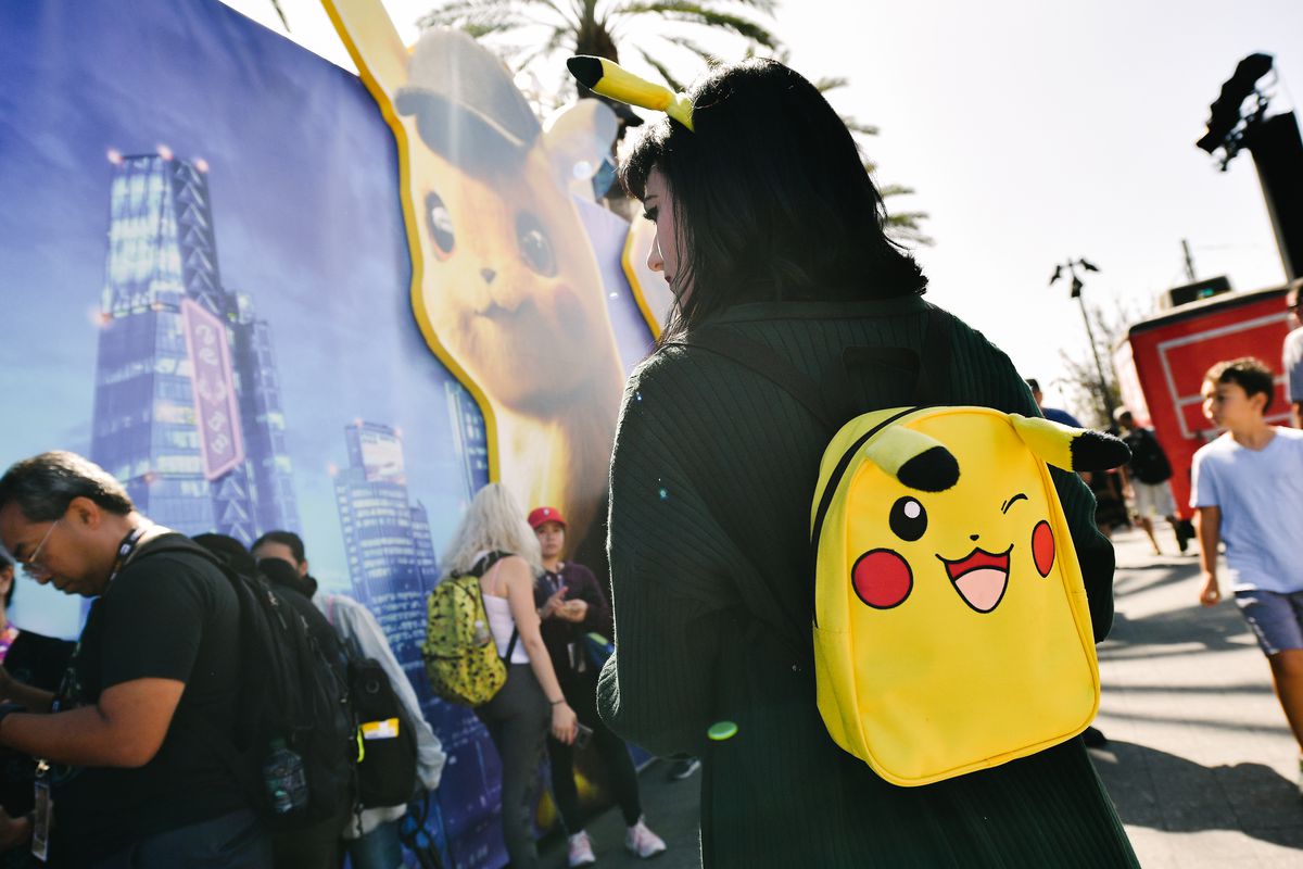Photo shows a woman wearing a yellow Pikachu backpack in the foreground, in the background many people are waiting in a line.