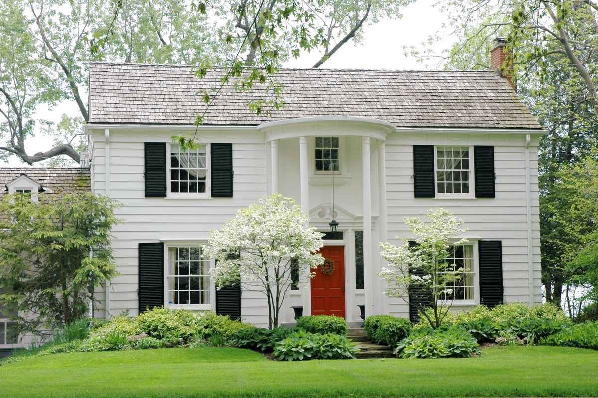 A white colonial house with red front door, black shutters, tall white columns, and bright green front yard. 