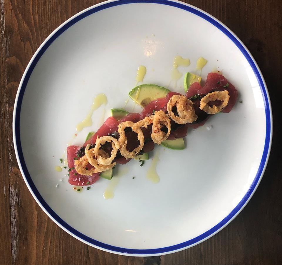 Several plump pieces of raw tuna are arranged in a line across a white plate with a blue border and garnished with several accoutrements, including a drizzle of oil.