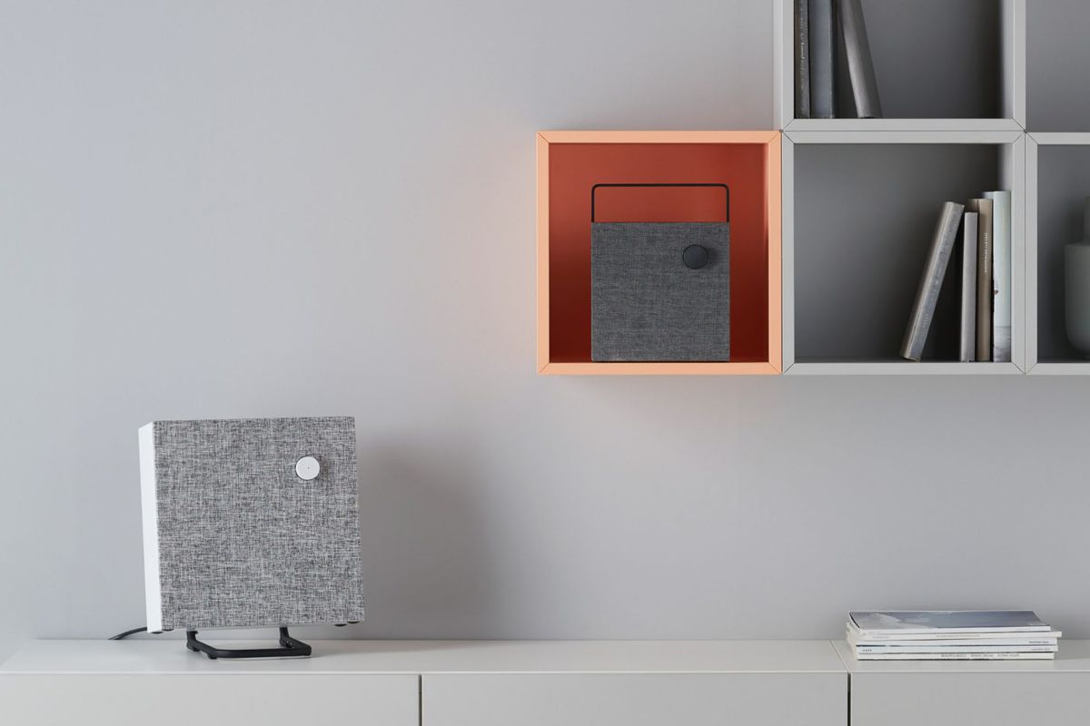 Flat box speaker on a stand on top of a dresser, and a smaller one placed inside a wall-mounted square shelf. 
