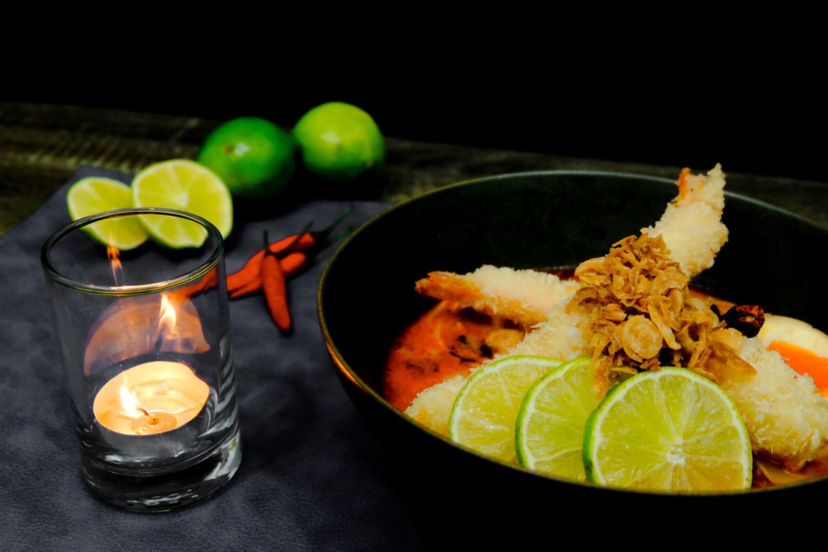 Two tempura shrimp stick out of a black bowl of reddish broth, garnished with sliced limes and crispy shallots.