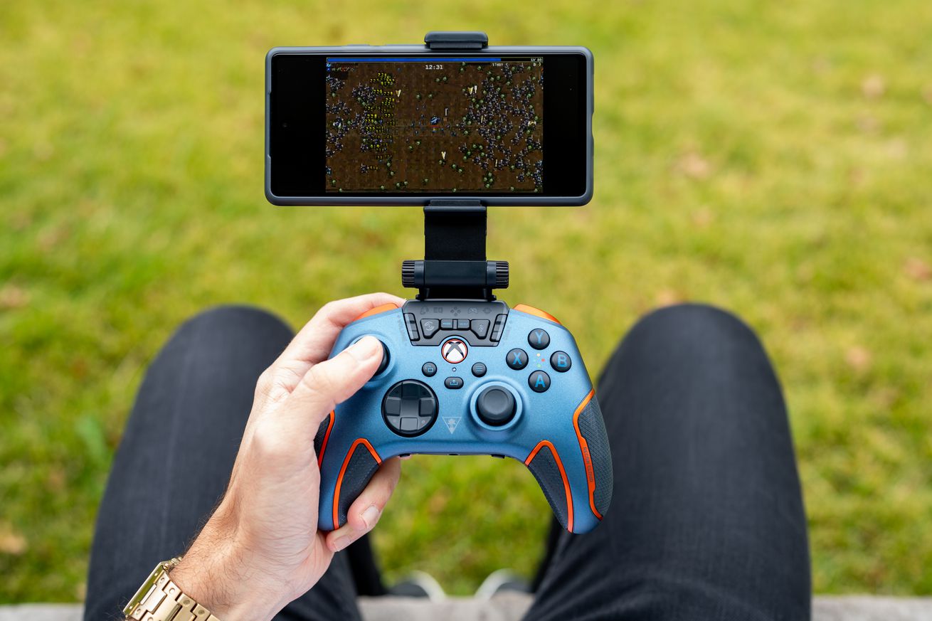 A person holding the blue and orange Turtle Beach Recon Cloud controller above their lap, outside in front of green grass. The controller has a smartphone mounted to its top, streaming a game from the cloud.