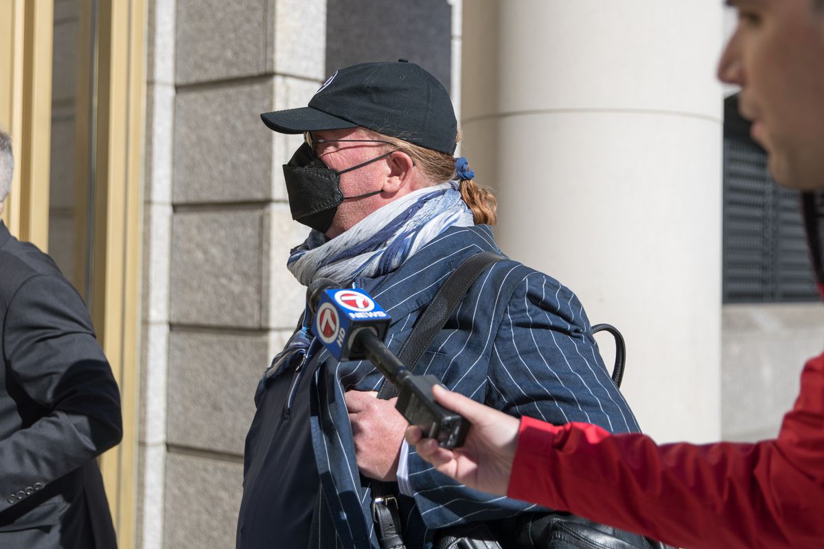 Man wearing baseball cap and mask walks past the microphone of a reporter.