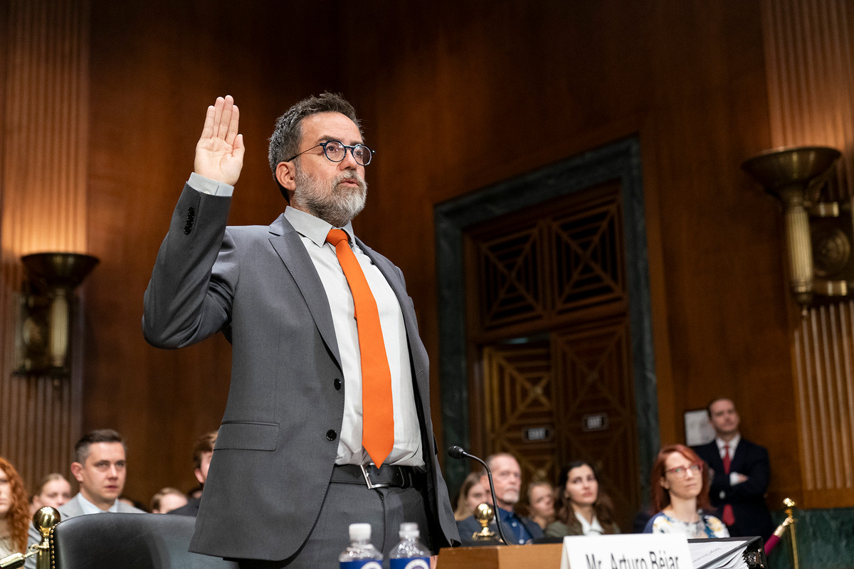 Arturo Bejar, wearing a gray suit with a white shirt and an orange tie, raises his right hand while standing in a Senate subcommittee hearing.