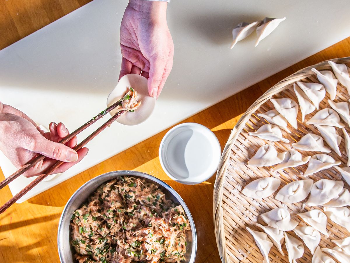 A pair of hands places filling inside a dumpling skin beside a bowl of filling and platter of completed dumplings.