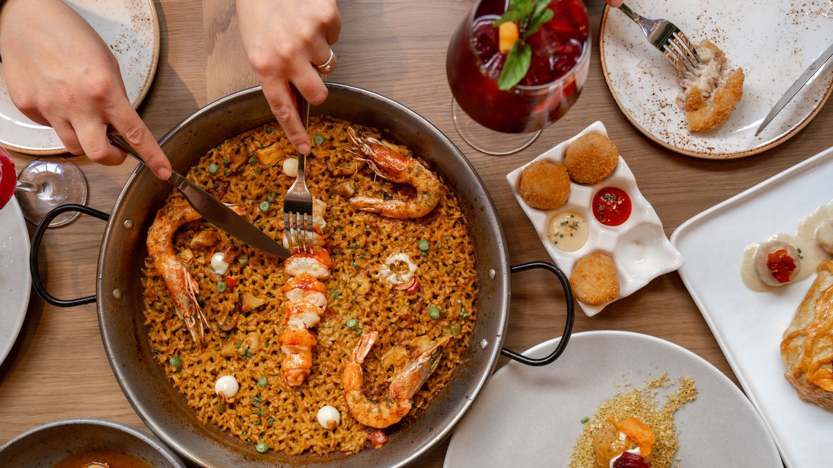 Hands cut into a seafood paella on a light wooden table with surrounding snacks, at a daytime restaurant.