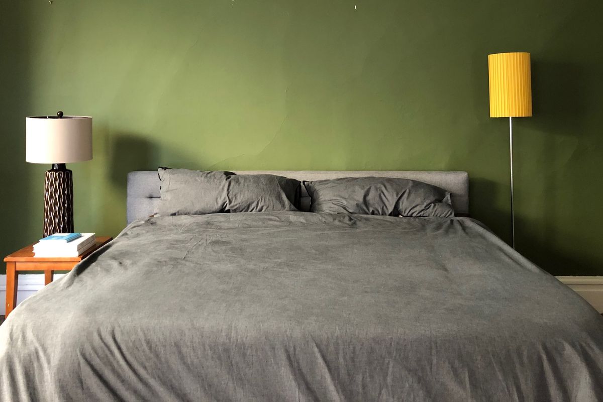 A large bed with gray duvet, a dark green wall, a wooden nightstand with a lamp and three books.