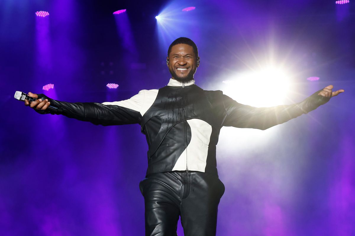 A photo of Usher at 2023 The Roots Picnic. He’s wearing a black and white leather outfit and is on stage performing. He’s standing smiling with his palms and arms outstretched. 
