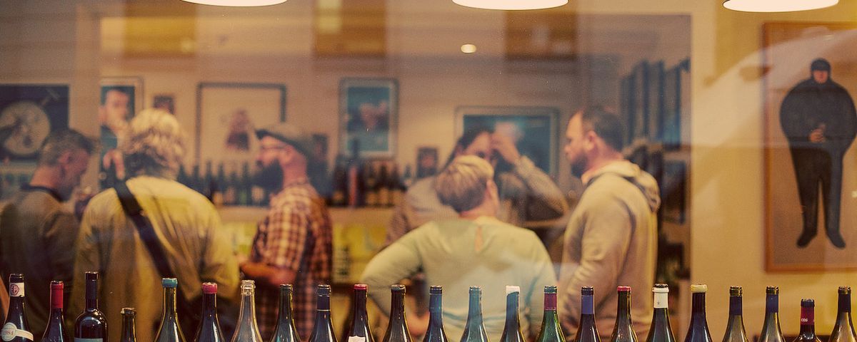 A crowd of wine drinkers seen through a front window, its sill lined with emptied bottles.