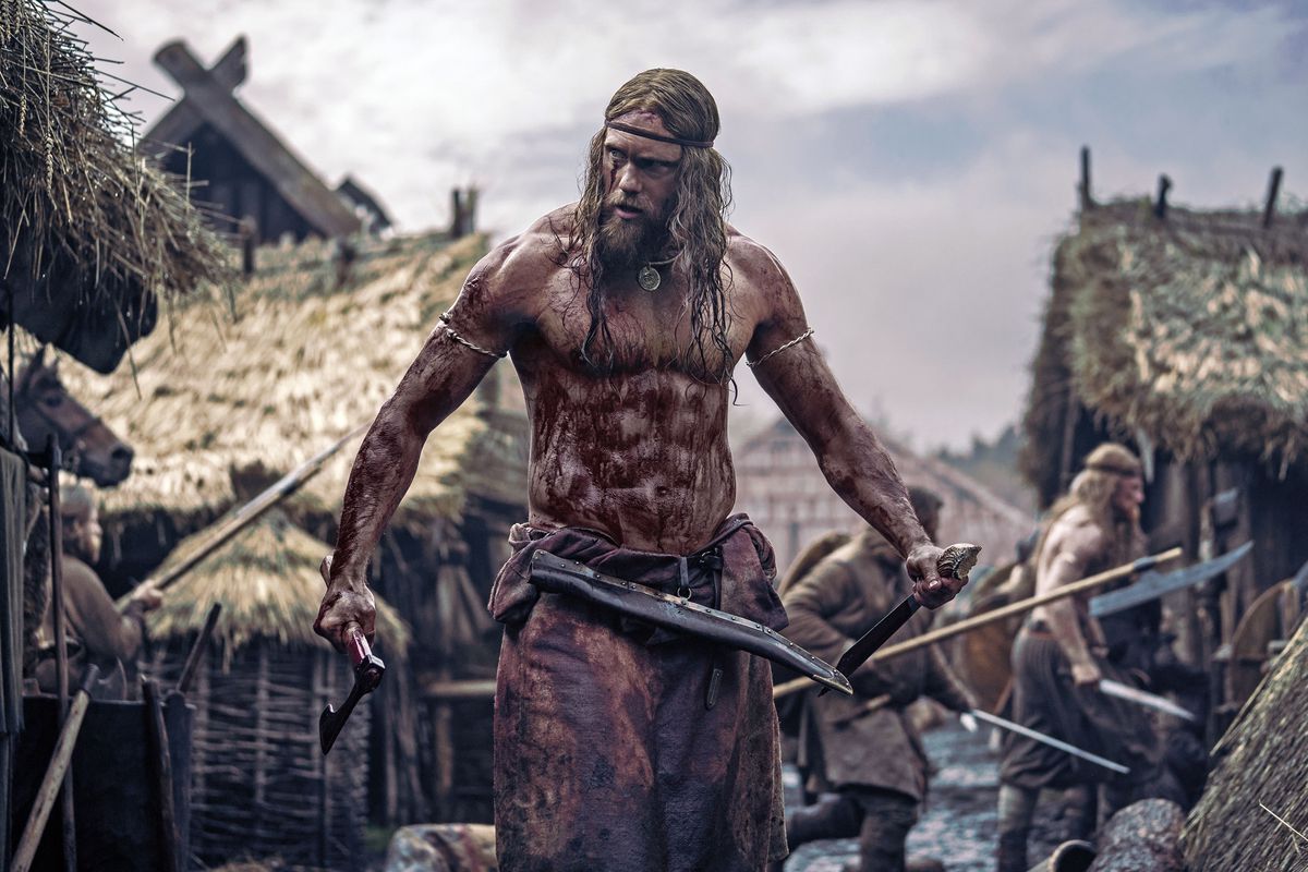 A Viking, half-naked and covered in blood, stands in the midst of a village.