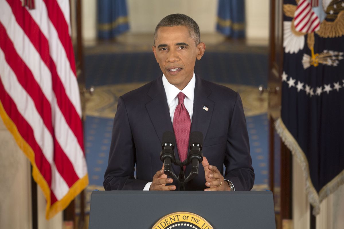 Barack Obama standing at a podium, flanked by American flags on either side.