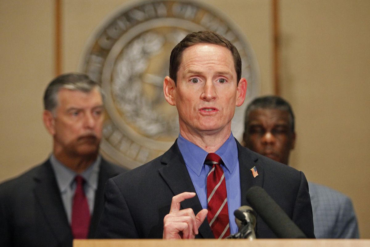 Dallas County judge Clay Jenkins, wearing a grey suit, blue shirt, and red tie, standing at a podium.
