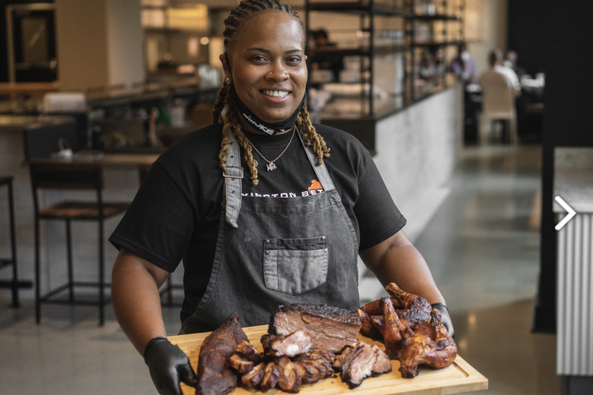 A woman smiles and holds a large cutting board full of barbecue meats.