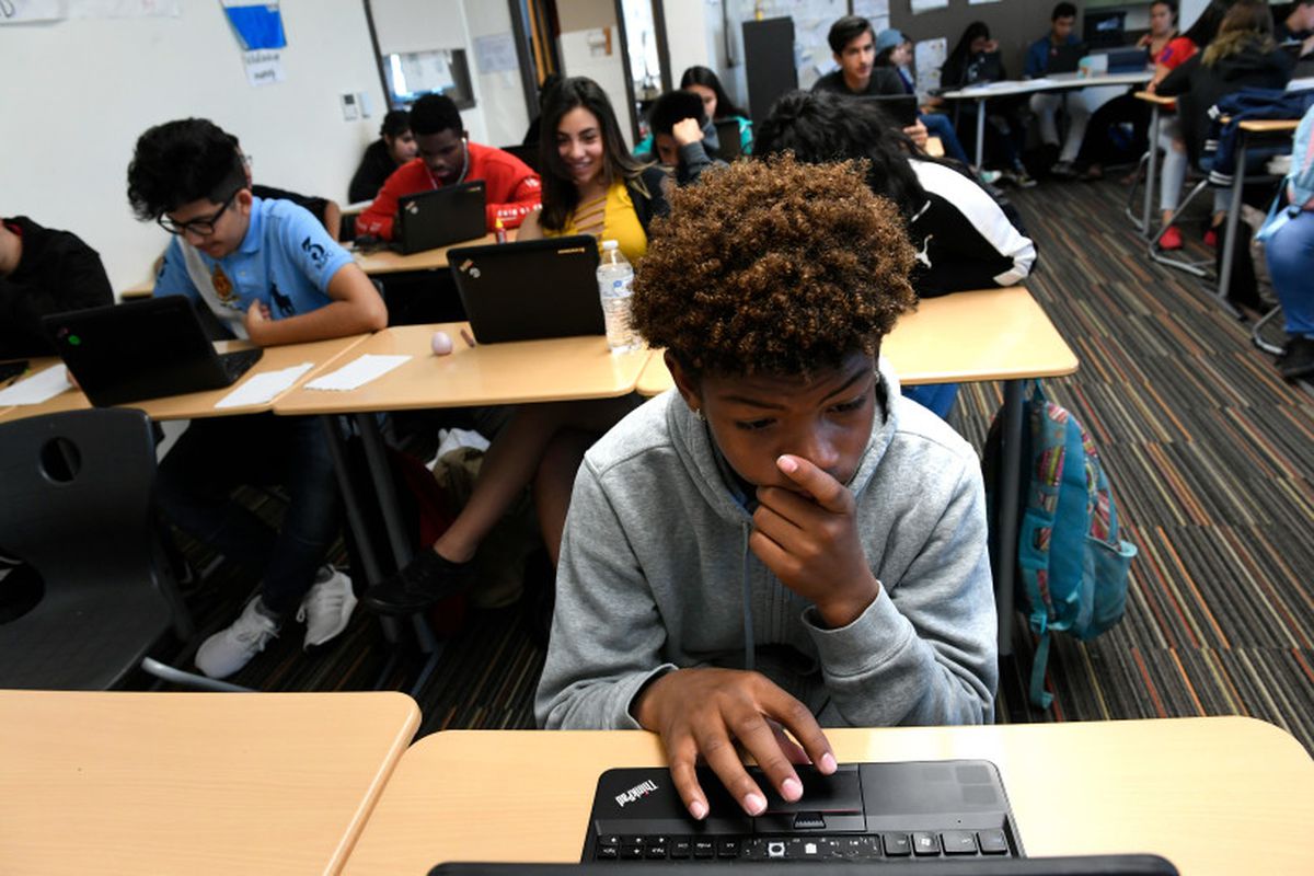 A high school student works at a computer amongst his classmates, several of which are behind him.