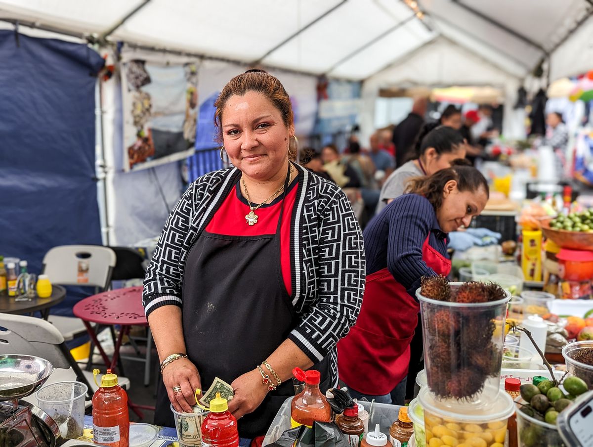 A woman in red shirt and black apron smiles at the camera with a delicate gold chain on as she handles cash from across a table at a street food vendor market in Los Angeles.