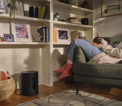 A Sonos Sub Mini subwoofer in a living room with a person relaxing on a couch nearby.