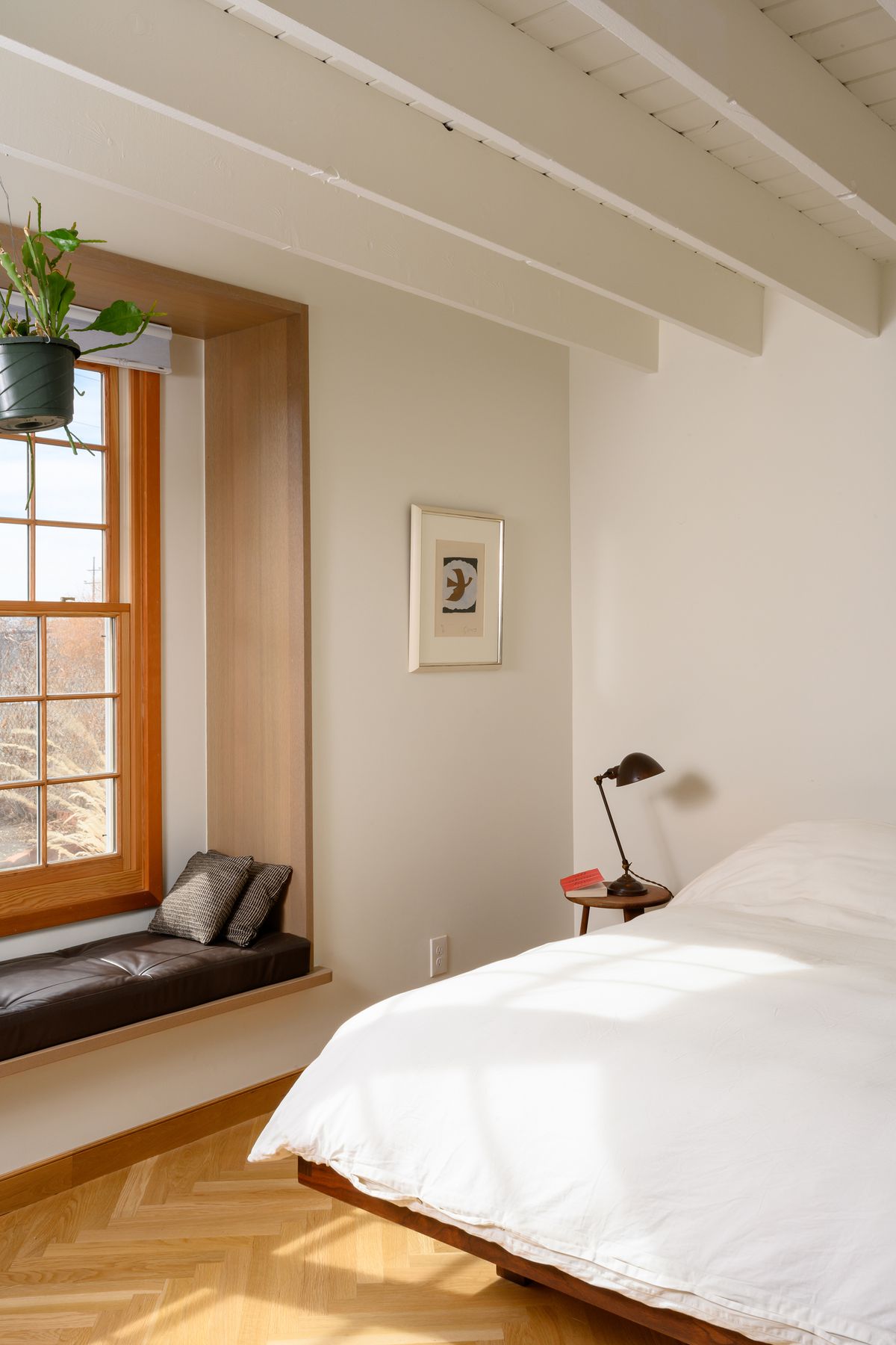 The bedroom, ceiling beams are painted white, there is a herringbone wood floor, a white bed spread and a window seat with a brown leather cushion and plant hanging.