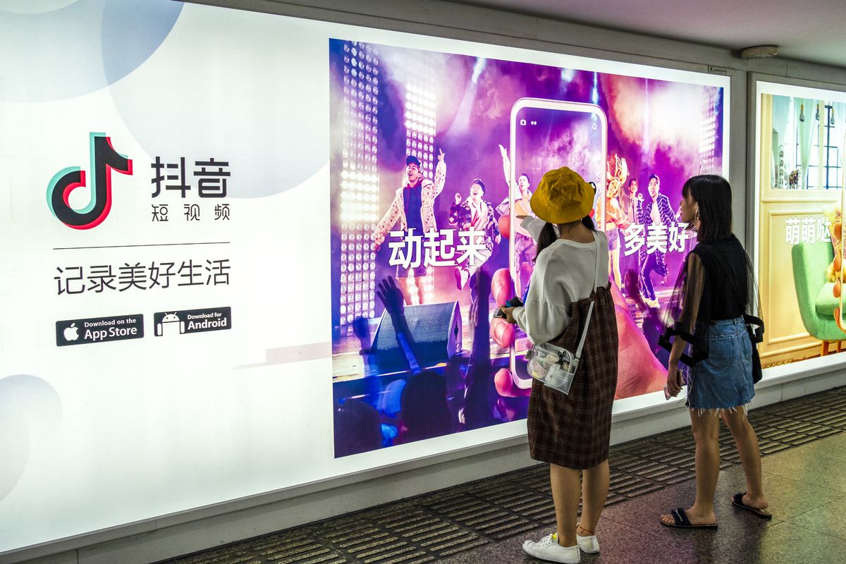 Two young women stand in front of a large digital billboard watching it display a Chinese ad for TikTok.