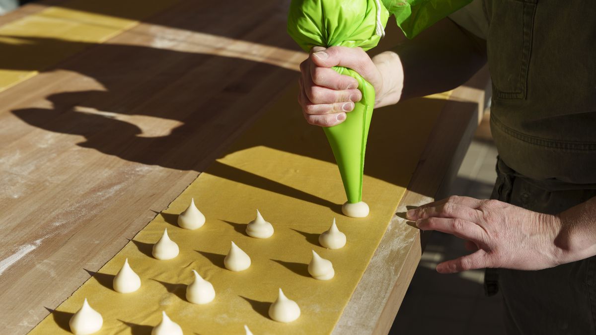 A long sheet of pasta on a wooden table; a person’s hands piping white parmesan filling in a green plastic bag onto it.
