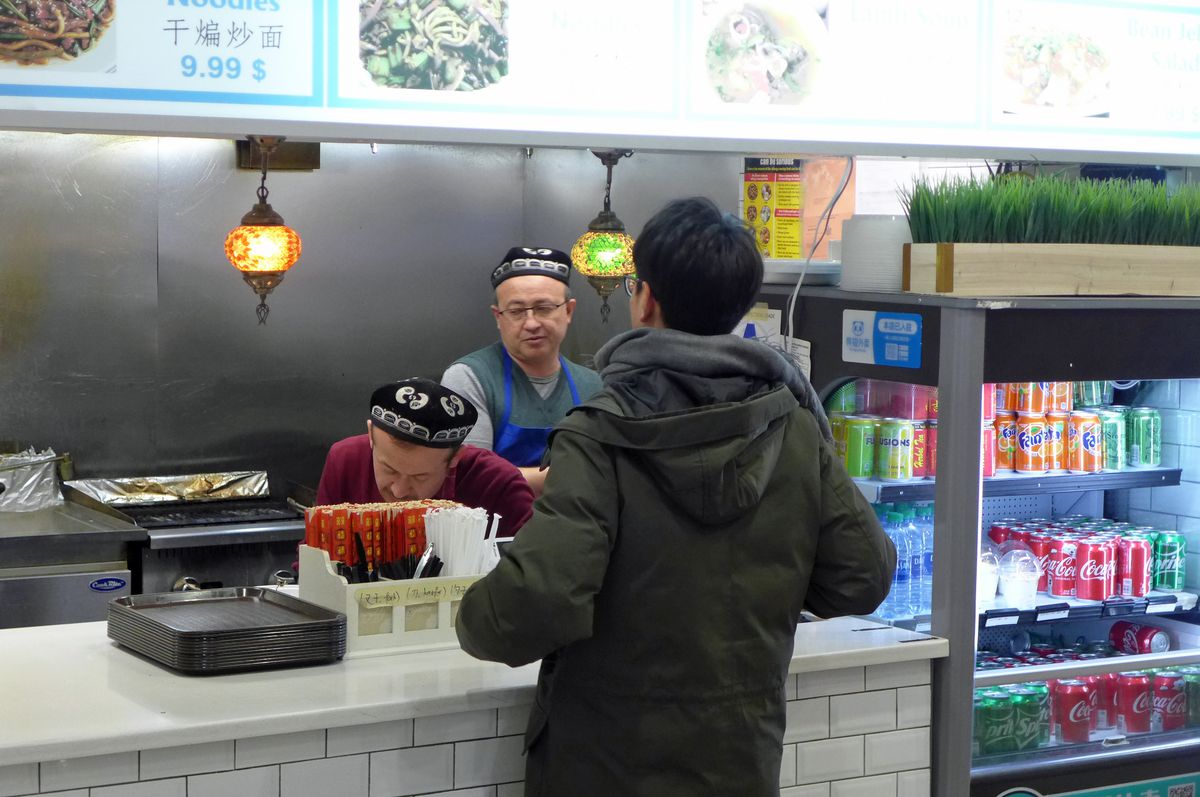 A customer stands before a counter behind which two men in blue skullcaps fill his order.