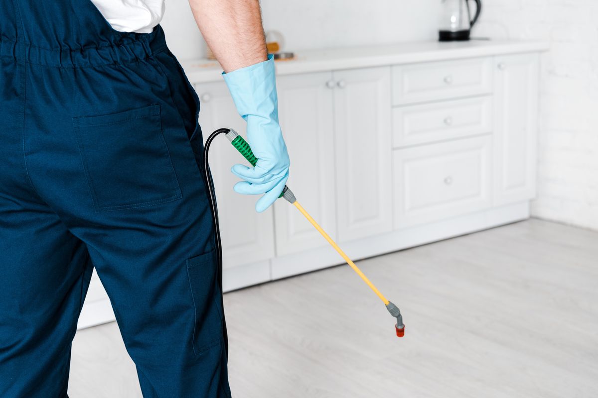 A pest control specialist wearing a navy blue jumpsuit holds a yellow wand in a white kitchen of a home with gray floors.