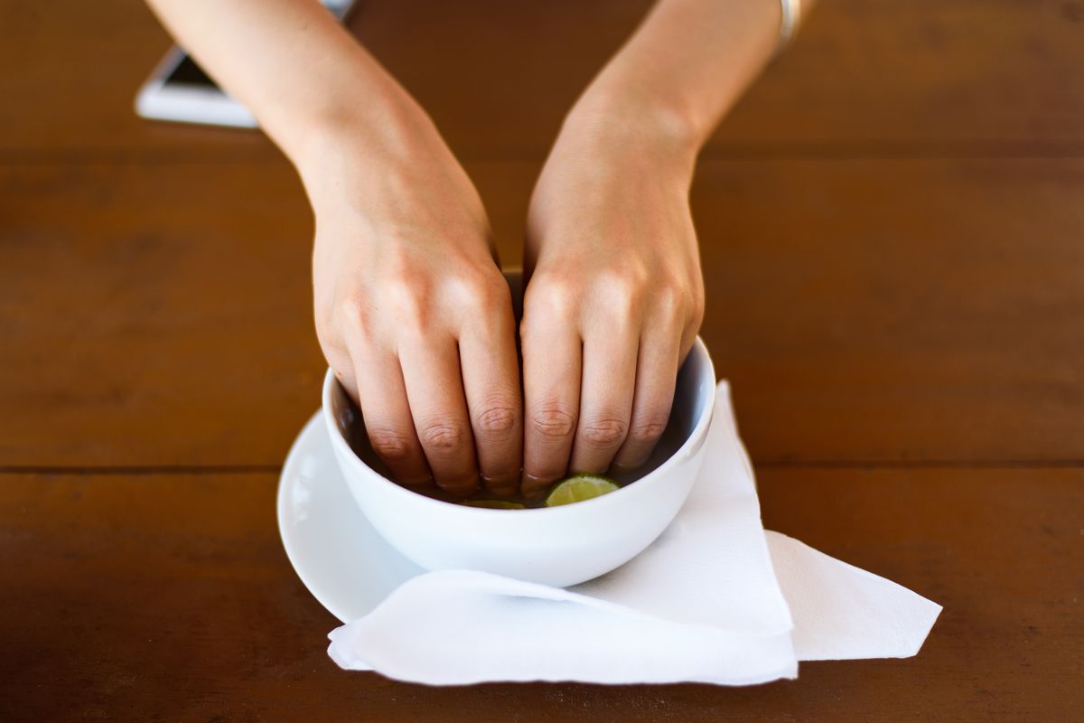 Two hands dipping into a bowl containing water and a wedge of lime.