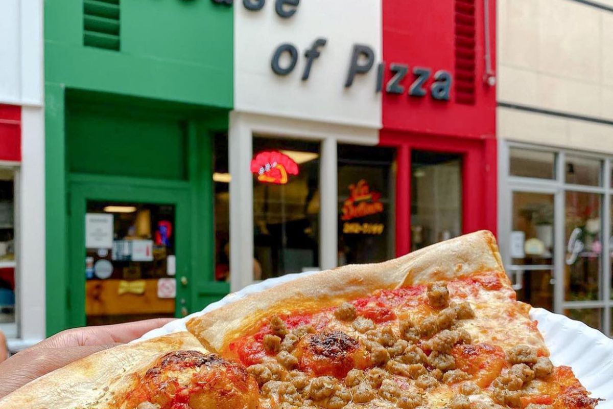 two slices of sausage pizza on a white paper plate being held up against a red, white, and green storefront that says “House of Pizza”