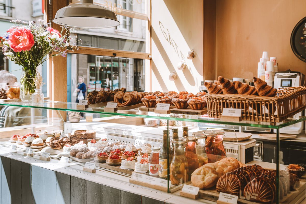 A sunny pastry counter, with a basket of croissants, rows of pastries, bottles of sweet drinks, and a bouquet of flowers. 
