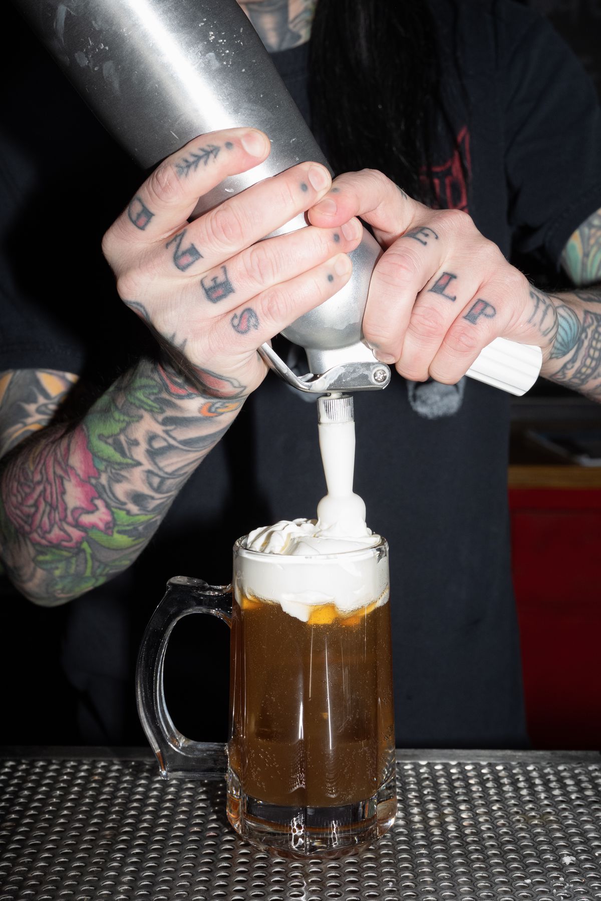 A person squeezing white foam on top of an amber colored cocktail in a rootbeer mug.