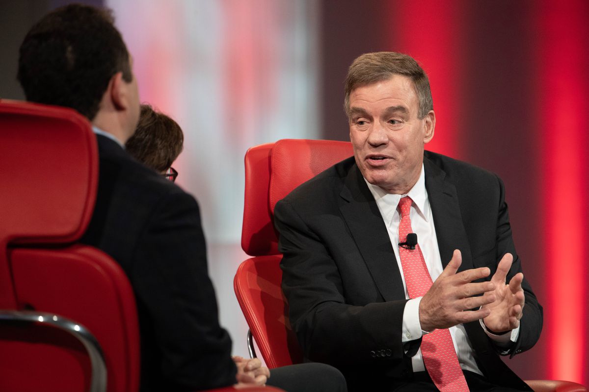 US Senator Mark Warner speaking onstage while seated in a red chair.