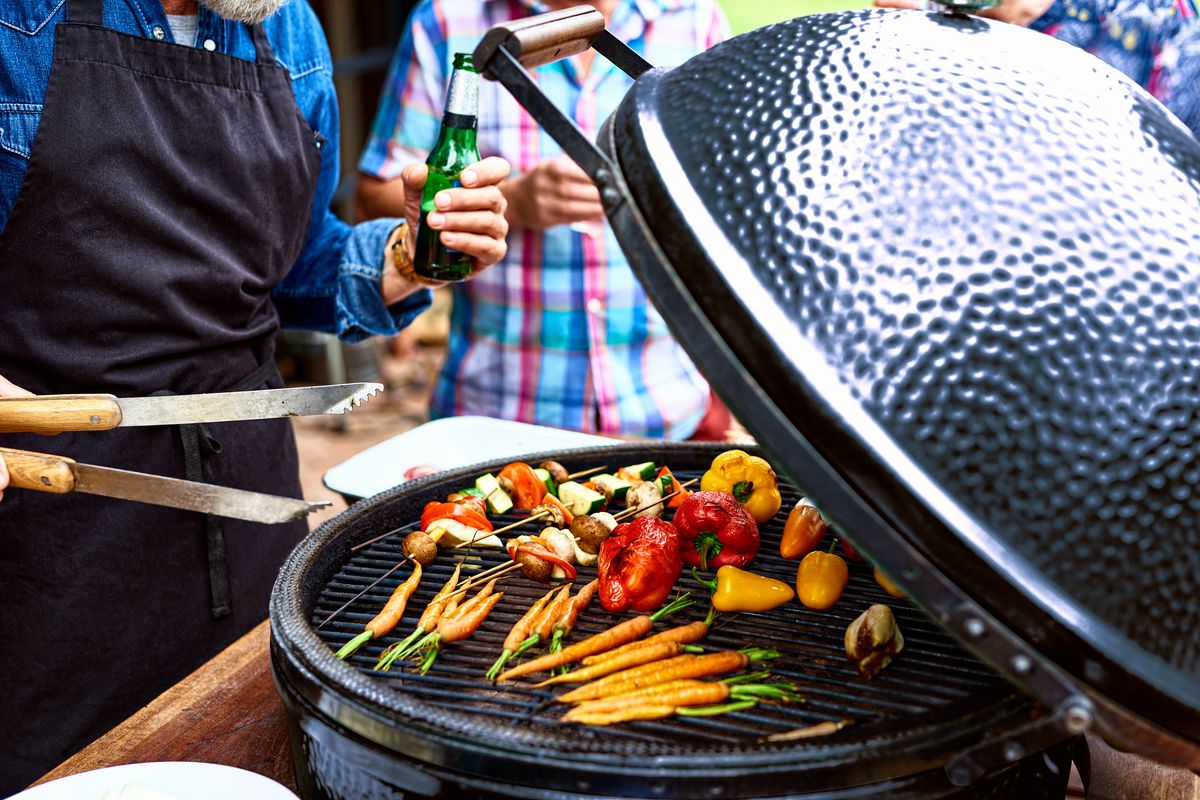 An array of vegetables is shown grilling on a kettle-style charcoal grill; two people stand next to it, one holding a pair of tongs.