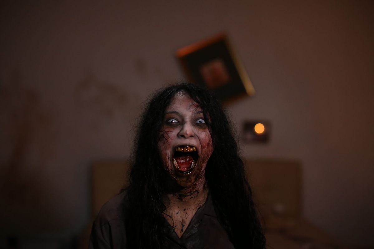 A woman with long dark hair, rotting teeth, and white skin with deep red veins screaming in a disheveled room in Blood Flower.