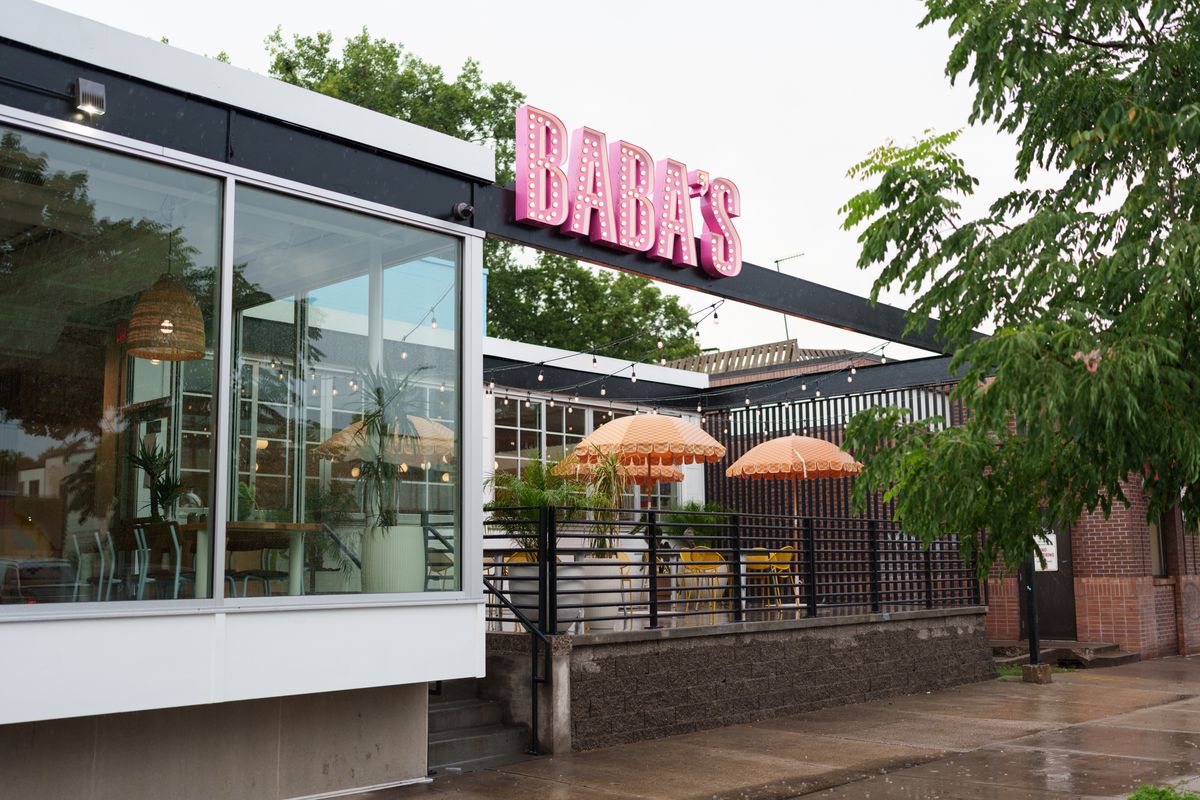 The exterior of a restaurant with big windows and a pink sign that says “Baba’s” above a patio. 
