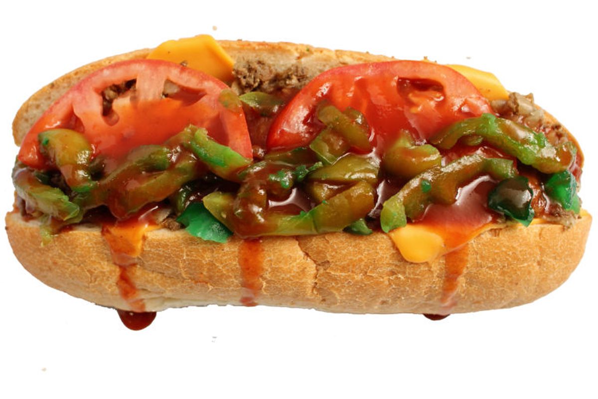 A steak sandwich on a long hoagie roll topped with sweet red sauce, two tomato slices, green peppers, and sliced yellow cheese.