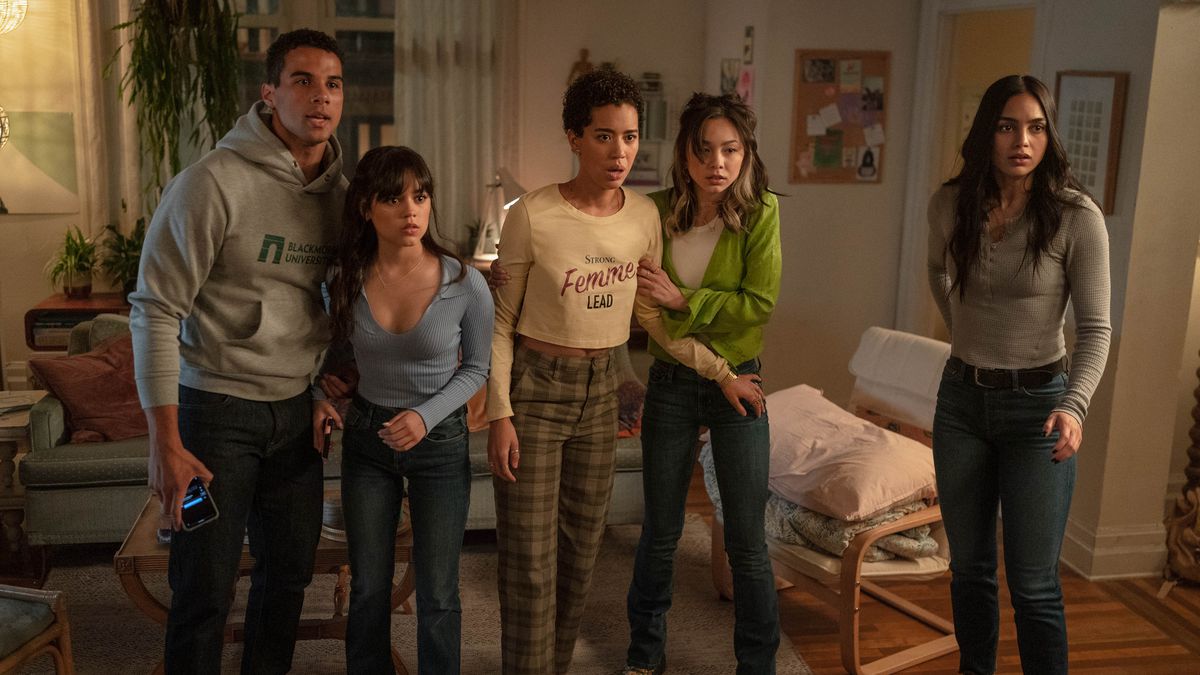 Five cast members from Scream VI (Devyn Nekoda, Jasmin Savoy Brown, Jenna Ortega, Mason Gooding, Melissa Barrera) stand together in a living room, clutching each other for support, and stare at something offscreen in Scream VI