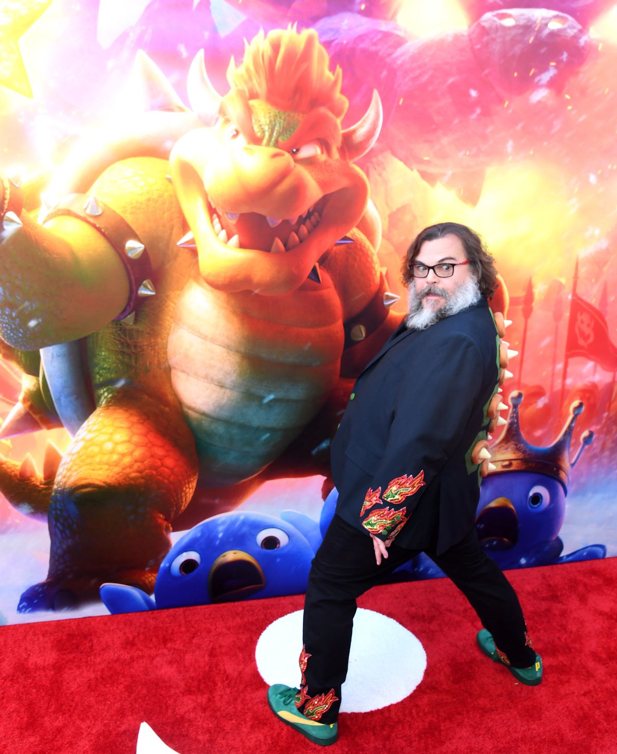 Jack Black poses on the red carpet at the LA premiere of The Super Mario Bros. Movie. He is wearing a suit with Bowser’s back spikes on it, with flame embroidery on the suit and pant cuffs.