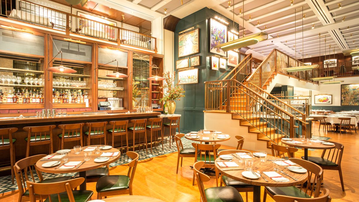 Union Square Cafe’s dining room has a wooden bar, wooden chairs with dark leather seats, and stairs that go up toward another level