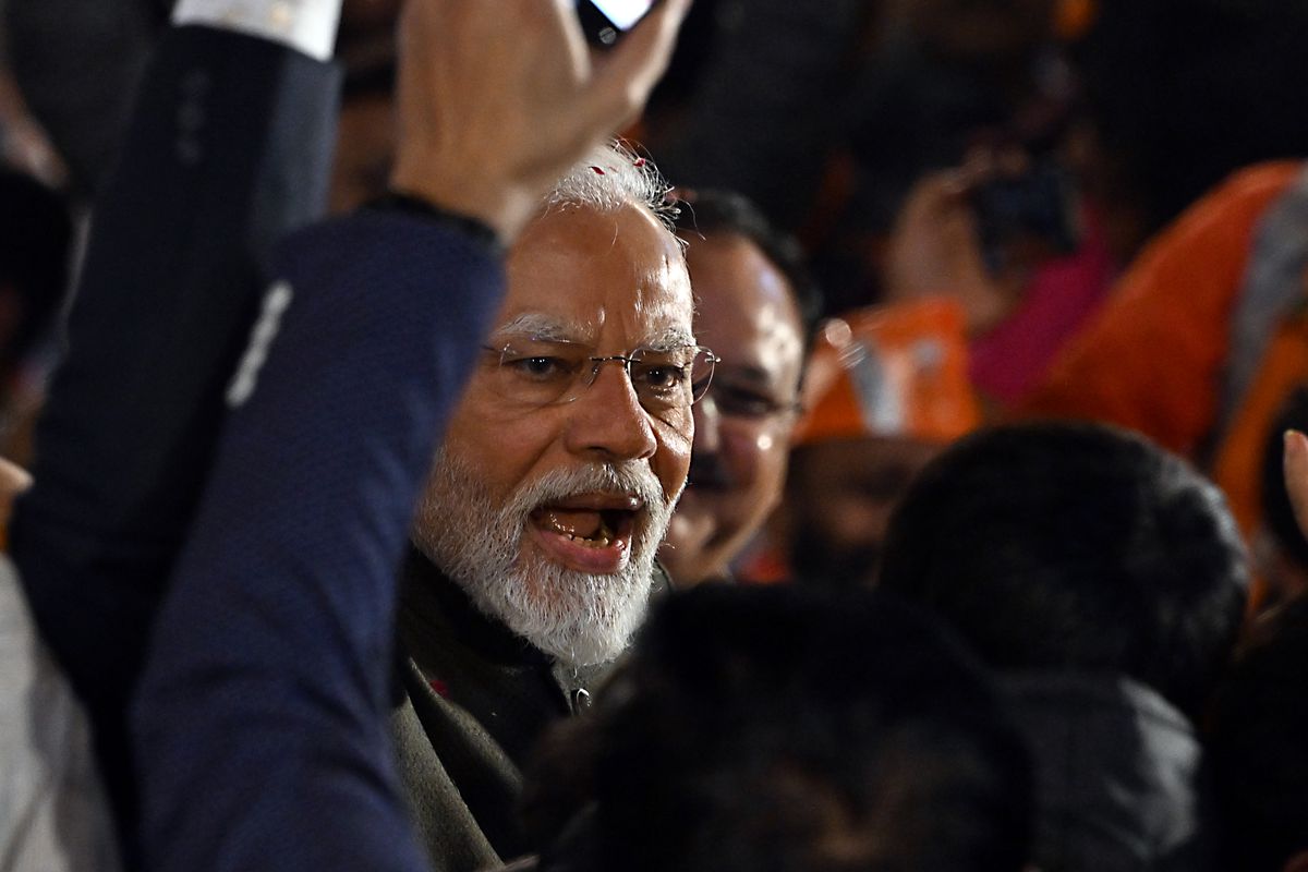 Narendra Modi, wearing frameless eyeglasses and a white beard, pictured mid-speech, lifting an arm in the air.