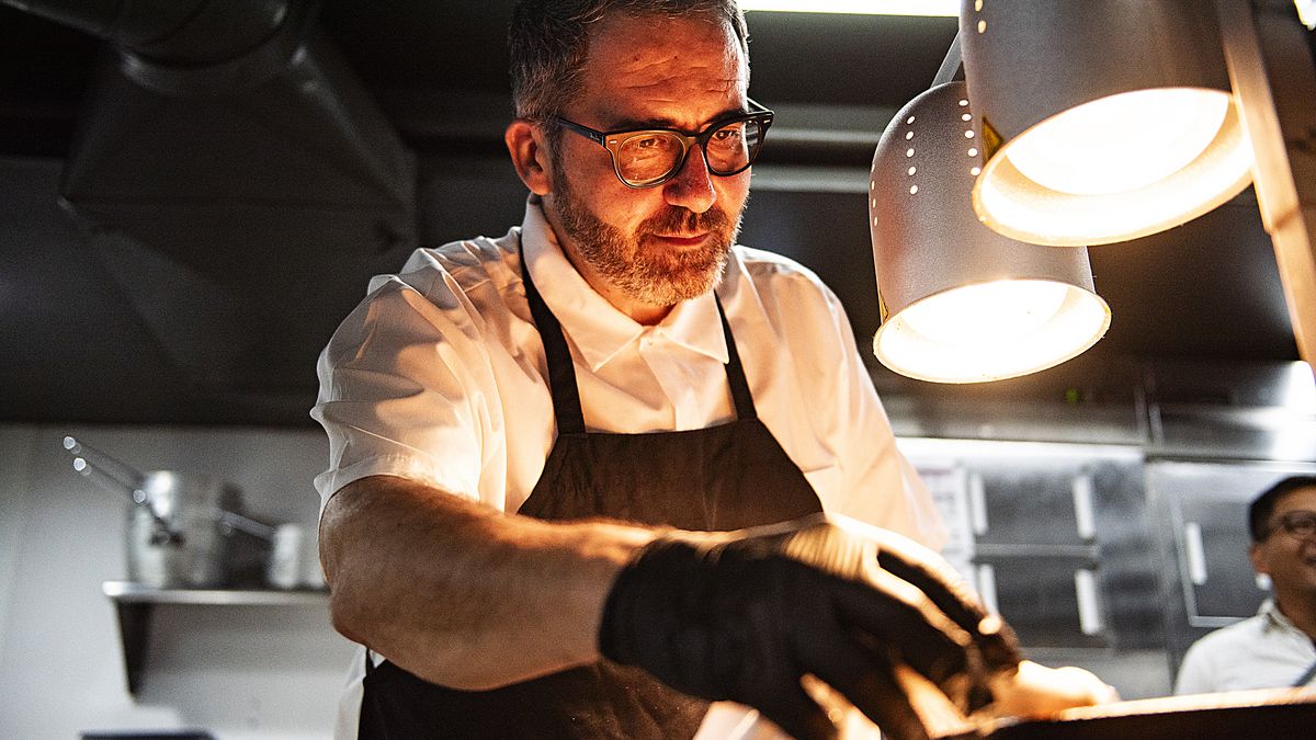 Chef Raúl Cob of Aventura in Ann Arbor, Michigan making paella, wearing black-rimmed glasses, a white jacket, black apron, black gloves, standing above two lamps and reaching froward.