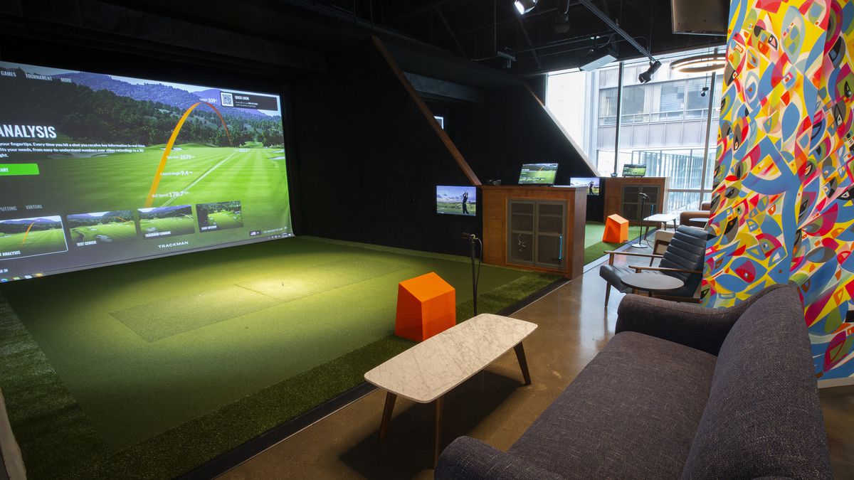 A row of golf simulator bays with large screens, putting greens, and lounge seating.
