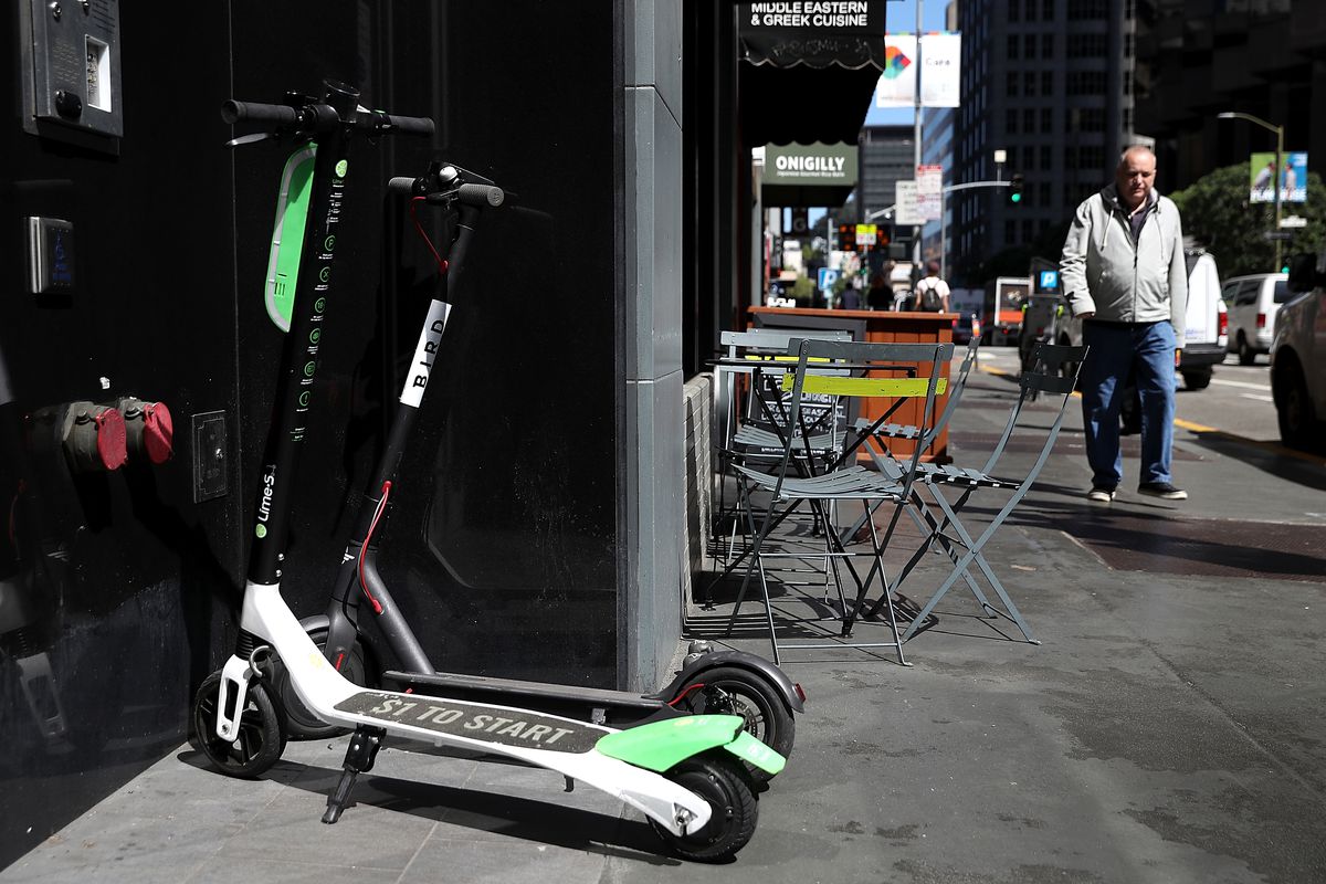 Green and white Lime scooters parked on the sidewalk. A male pedestrian walks near them.