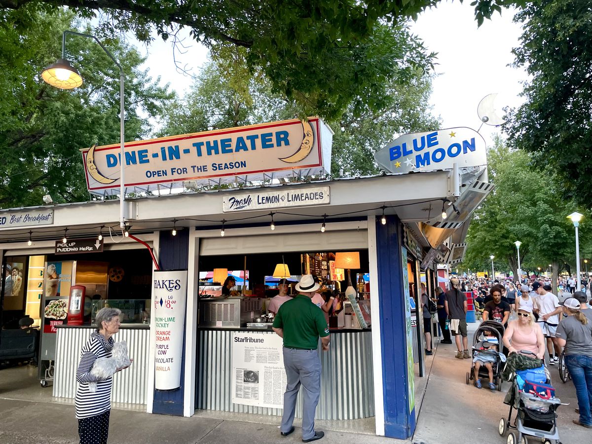 A building with signs that say “Dine-in-Theater” and “Blue Moon” in a canopy of trees with people all around. 