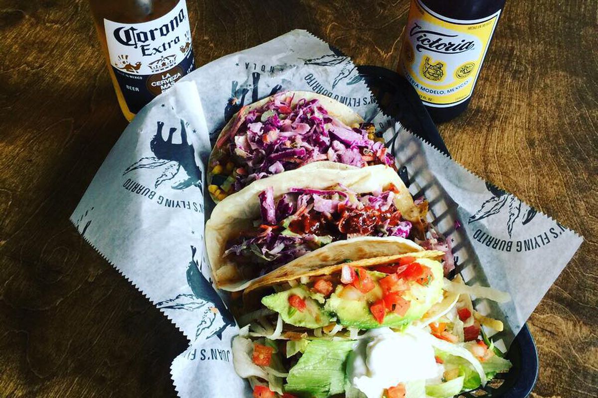 A platter of four tacos, two topped with red cabbage and two with tomatoes and iceberg lettuce, on a table with two bottles of beer.